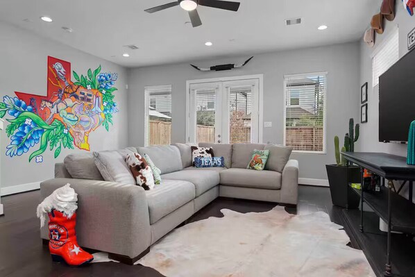 A modern living room with a gray sectional sofa, a cowhide rug, and a colorful mural on the wall. - Texas Cities Themed House, Hot tub. Don't Mess With Texas (Houston)