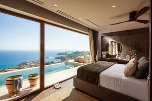 Ein Schlafzimmer mit großem Fenster und Blick auf die Küste, einem Bett, einem Deckenventilator und einem Nachttisch mit Lampe.