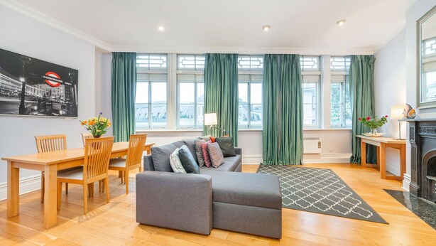 Ein modernes Wohnzimmer mit Sofa, Esstisch und Stühlen, großen Fenstern mit grünen Vorhängen sowie einem Kamin.