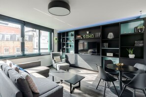 A modern living room with a sofa, armchairs, a coffee table, and a dining table with chairs. There is a bookshelf with decorative items and a sign that reads "SOHO". - The Slaycation Crib - Soho - by Frankie Says (London)