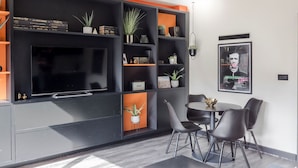 A modern living room with a large flat-screen TV, built-in shelving with books and plants, a dining area with a round table and chairs, and a framed picture on the wall. - The Peaches Cream - Soho - by Frankie Says (London)