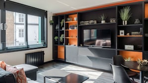 A modern living room with a large window, a sofa, a coffee table, and built-in shelving with decorative items. - The Peaches Cream - Soho - by Frankie Says (London)