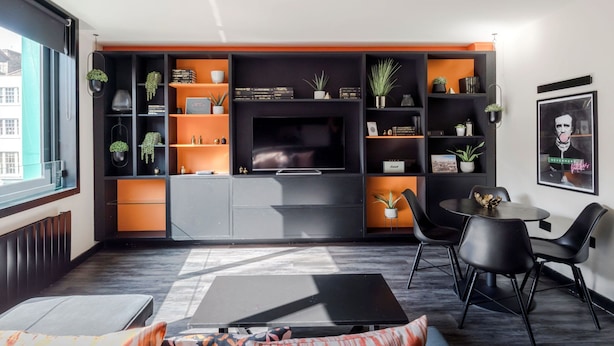 A modern living room with a large flat-screen TV, black and orange shelving, a dining area with black chairs, and a window providing a view of the city. - The Peaches Cream - Soho - by Frankie Says (London)
