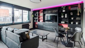 Una sala de estar moderna con sofá, mesa de centro y comedor. Hay un ventanal grande, un televisor y estanterías con plantas y libros.