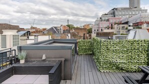 Una terraza en la azotea con piso de madera, un seto verde y vistas de edificios circundantes.