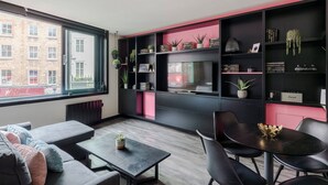 Een moderne woonkamer met een bank, salontafel en een eethoek met stoelen. Er is een groot raam met uitzicht op een gebouw.