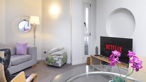 A modern living room with a sofa, ottoman, round mirror, lamp, TV, and a glass table with orchids. - The Aristocrat - Fitzrovia - by Frankie Says (London)