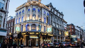 Een historisch gebouw met een opvallende hoekingang, een theaterscherm en een straatbeeld met voertuigen en voetgangers.