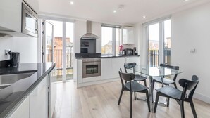 A modern kitchen with a dining area, featuring a glass table and chairs, stainless steel appliances, and large windows. - The Kings Residence - St Pancras - by Frankie Says (London)