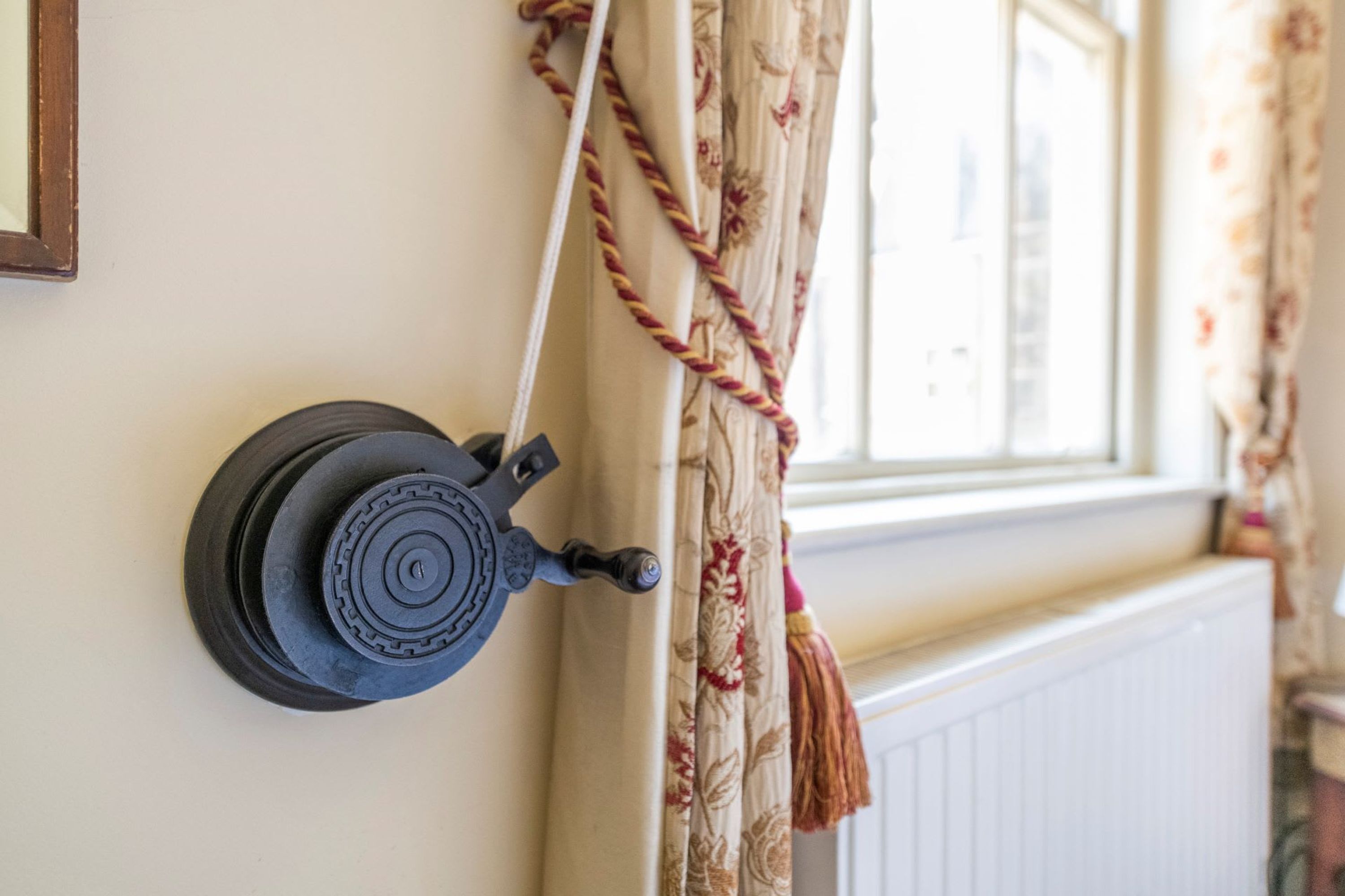 Un dispositivo fissato al muro, forse un termostato o un pannello di controllo, vicino a una finestra con tende floreali e un termosifone.