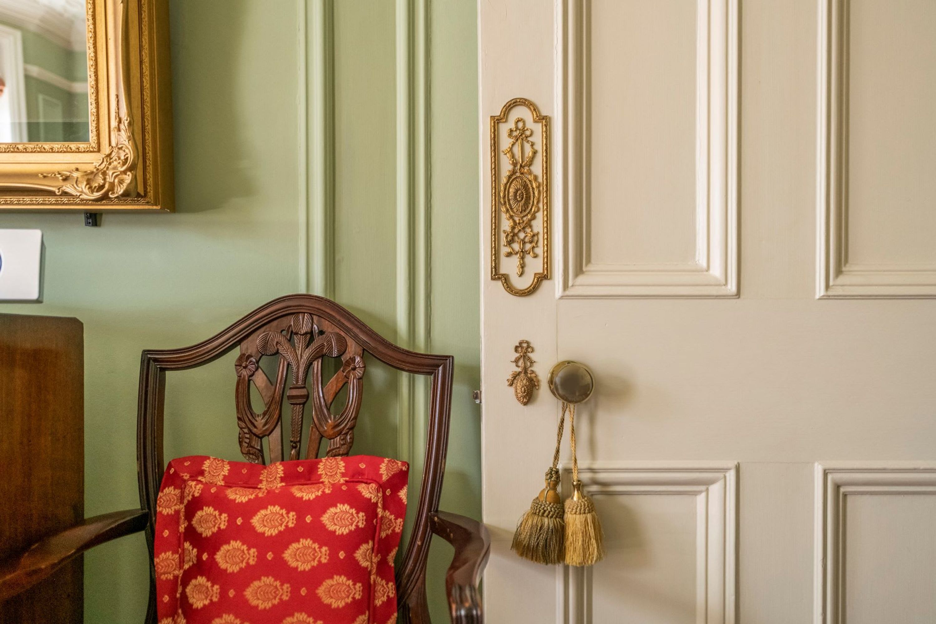 A room with a wooden chair, a door with a brass handle, and a mirror on the wall.