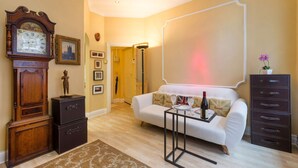 A living room with a white sofa, a side table with a bottle and glasses, a large clock, and a cabinet with drawers. - The Vintage - Fitzrovia - by Frankie Says (London)