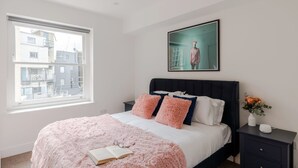 Un dormitorio con cama, mesita de noche, ventana y un cuadro en la pared.