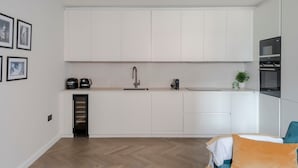 Une cuisine moderne avec des armoires blanches, un four encastré et un four à micro-ondes. On y trouve un petit évier et une machine à café sur le plan de travail.