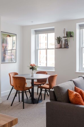 A dining area with a round table, six chairs, a vase with flowers, and a sofa with cushions.