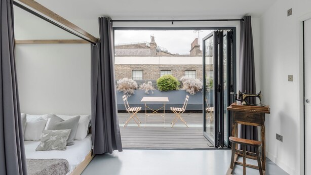Une chambre à coucher avec un lit, un tabouret en bois, une machine à coudre et une vue sur le balcon extérieur.