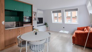 A modern kitchen with wooden cabinets, a dining area with a white table and chairs, a sofa, and a window with a view of buildings. - Casa Emilio - Soho - by Frankie Says (London)