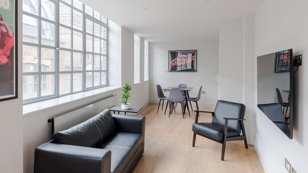 Una sala de estar moderna con un sofá de cuero negro, una mesa de comedor con sillas y un ventanal que proporciona luz natural.