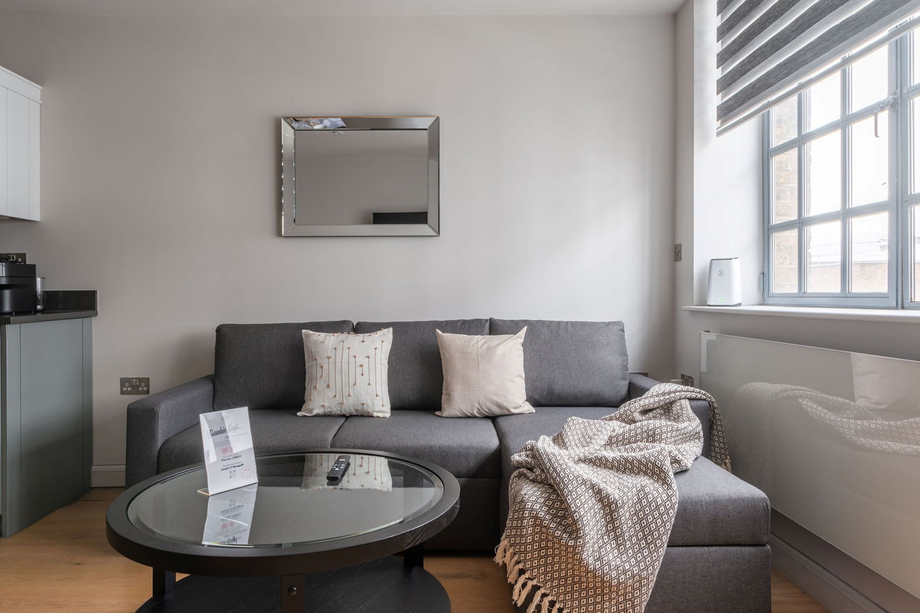 A modern living room with a grey sofa, a glass coffee table, and a large window with blinds.