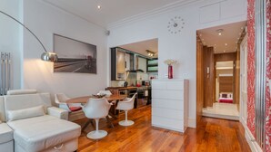 A modern living room with a sofa, dining table, and chairs. There is a kitchen in the background with a refrigerator and a microwave. - The Red Room Apartment - Fitzrovia - by Frankie Says (London)