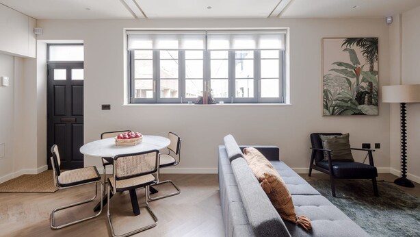 A modern living room with a grey sofa, a round dining table with four chairs, a black door, and a large window with blinds. - Parkside House - Regent s Park - by Frankie Says (London)