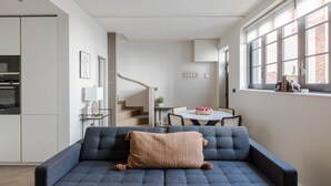 A modern living room with a dark blue sofa, a beige knitted throw pillow, and a dining area with a round table and chairs. - Parkside House - Regent s Park - by Frankie Says (London)