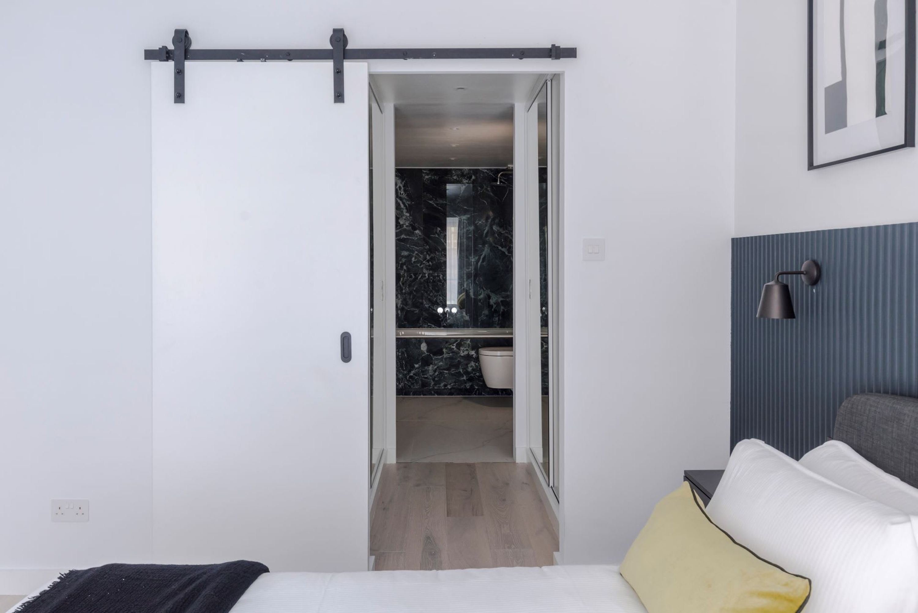A bedroom with a bed, a bedside table, a lamp, and a sliding door leading to a bathroom with a marble floor and a toilet.