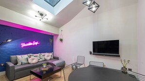 A modern living room with a neon sign, a flat-screen TV, a sofa, a coffee table, and a dining table. - The Insta Playhouse - Covent Garden - by Frankie Says (London)