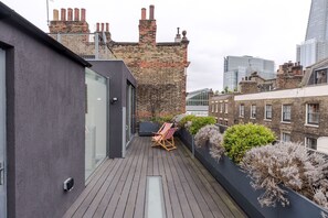 A modern rooftop terrace with a wooden deck, a single lounge chair, and a black planter with dried plants. - The Dream Factory - Borough - by Frankie Says (London)
