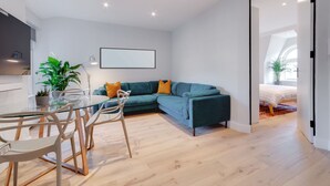 A modern living room with a glass dining table, teal sofa, and a large window. - Stairway To Heaven - Fitzrovia - by Frankie Says (London)