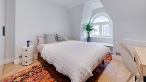 A bedroom with a large bed, a bedside table with drawers, a small potted plant, a window with a view, and a chair. - Stairway To Heaven - Fitzrovia - by Frankie Says (London)