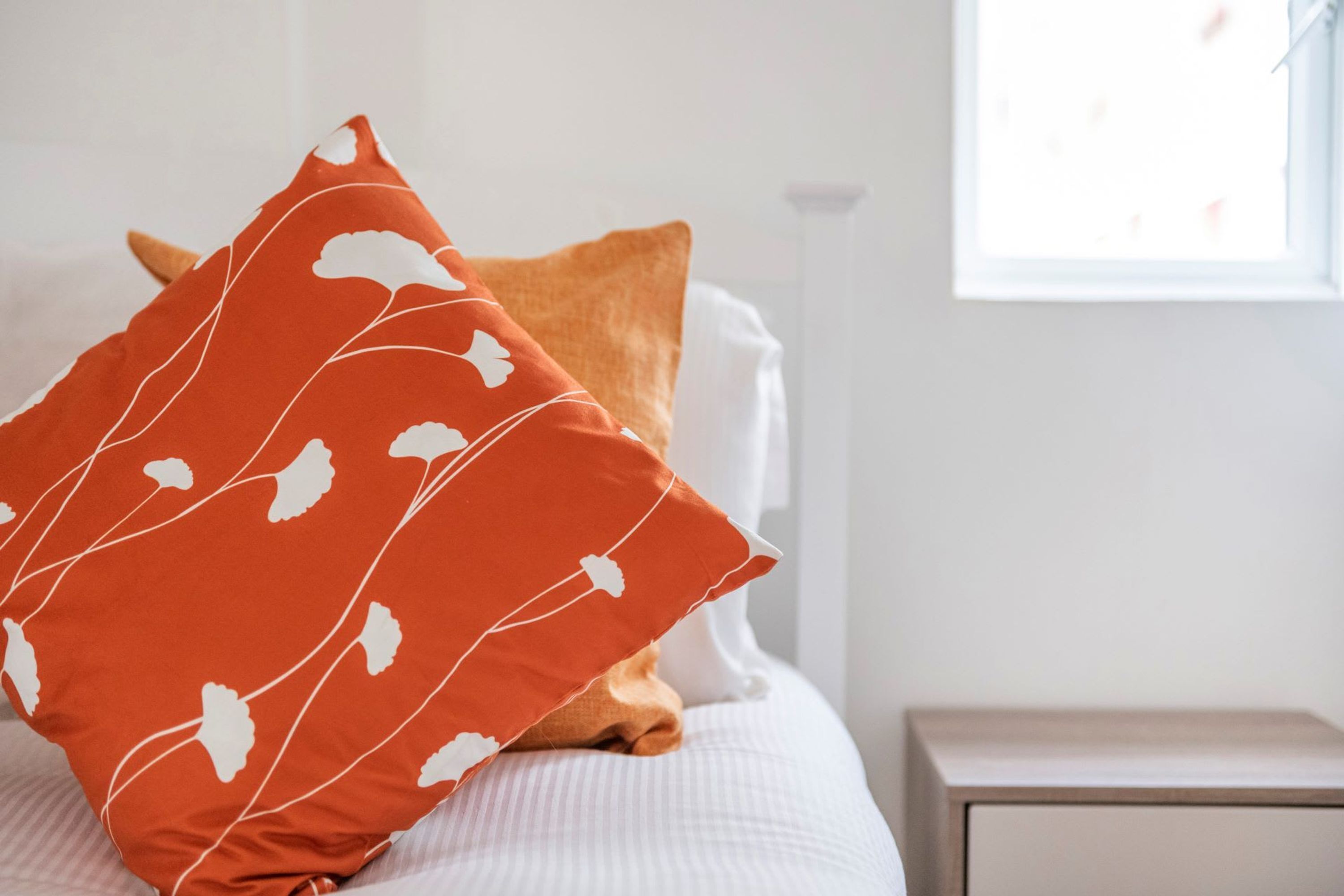 A neatly made bed with a white comforter and two pillows, one with a leaf pattern and another solid orange.