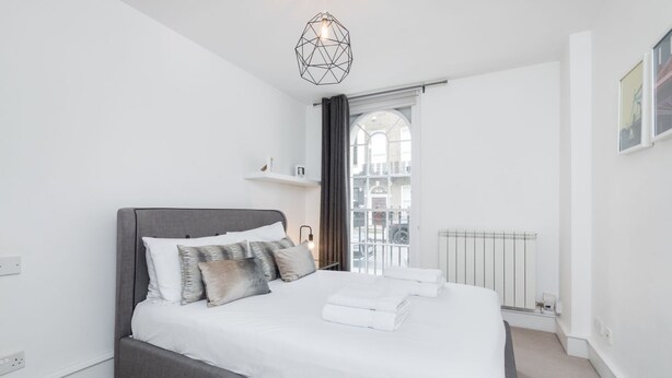 Une chambre moderne avec un grand lit, un luminaire suspendu aux formes géométriques, une vue par la fenêtre et un radiateur.