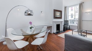 Un salon moderne avec une table à manger en verre, des chaises blanches, un canapé gris, une cheminée et une grande fenêtre avec des rideaux.