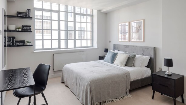 A modern bedroom with a large window, a bed with multiple pillows, a bedside table, a chair, and a small table with a lamp. - London s Calling - Farringdon - by Frankie Says (London)