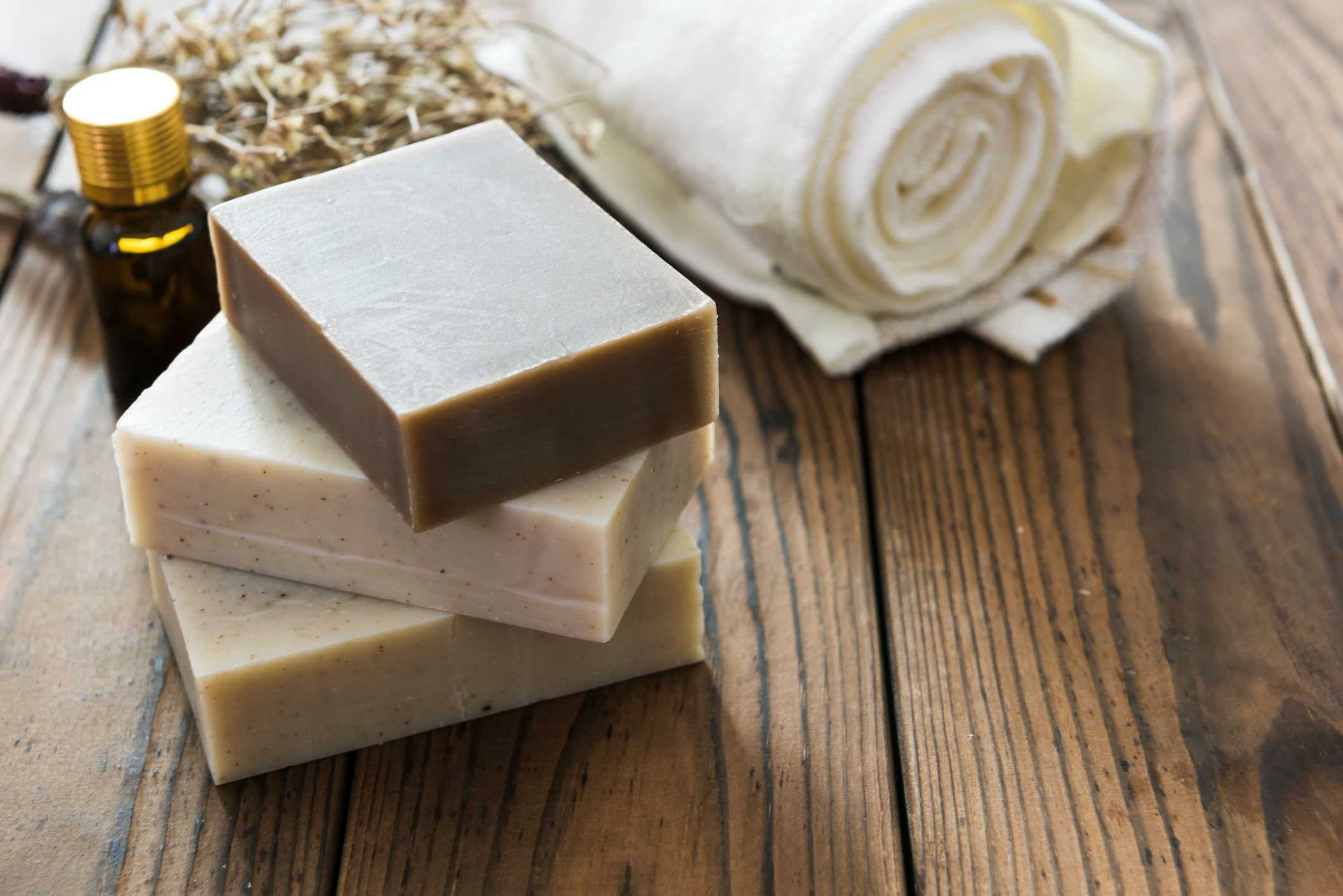 Three stacked bars of soap on a wooden surface with a rolled towel and a bottle in the background.