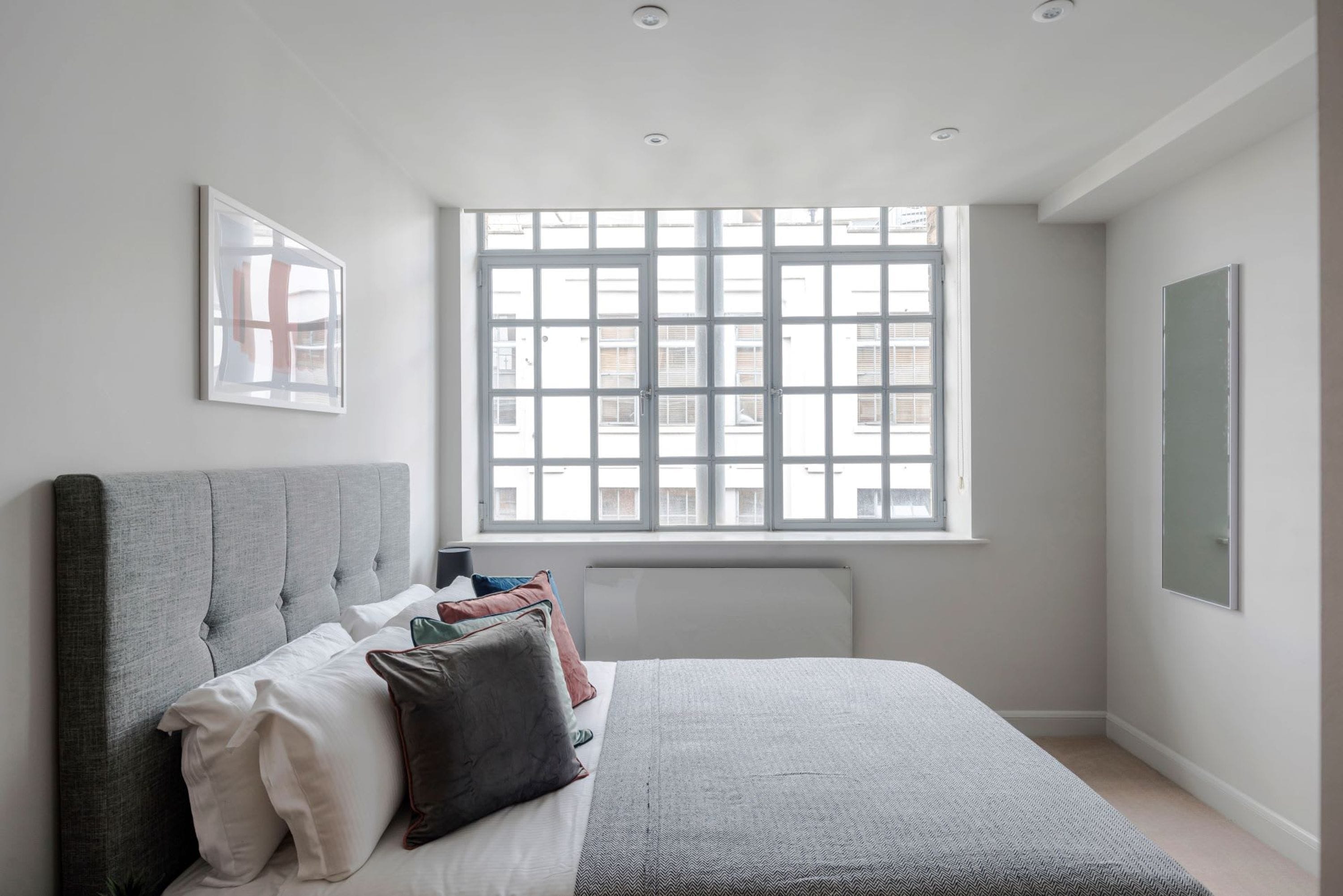 A bedroom with a large window, a bed with pillows, a radiator, and a framed picture on the wall.