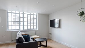 A modern living room with a sofa, a round coffee table, a TV mounted on the wall, and a large window providing natural light. - London s Calling - Farringdon - by Frankie Says (London)