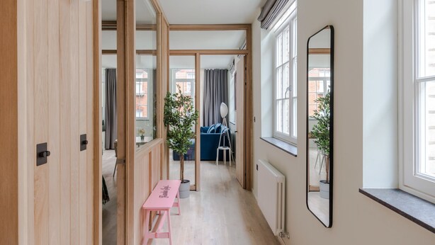 A hallway with wooden doors, a pink bench, a large mirror, and a view of a blue sofa and curtains in the background. - Casa Peppino - Soho - by Frankie Says (London)