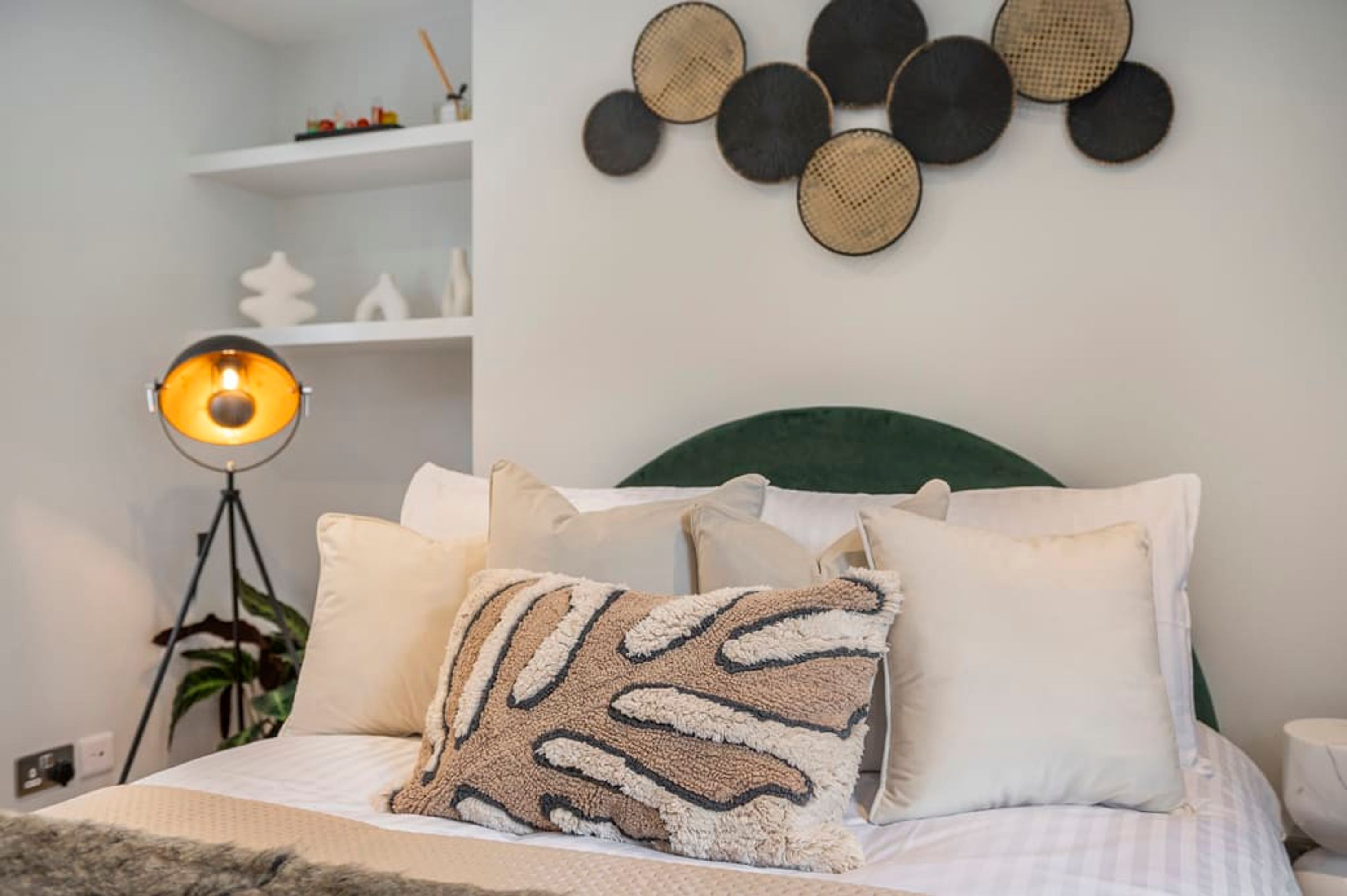 Un dormitorio ordenado con cama, almohadas, una lámpara de pie y un adorno en la pared.