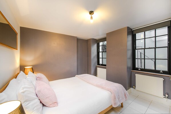 Un dormitorio con cama, lámpara de noche, ventana, radiador y vistas al exterior.
