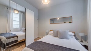 A modern hotel room with a large bed, a bedside table with a lamp, a mirror on the wall, and a window with curtains. - The Foxhole - Fitzrovia - by Frankie Says (London)