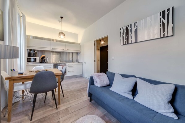 A modern living room with a blue sofa, a dining table with chairs, and a kitchen area in the background.