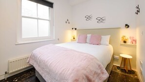 A bedroom with a large bed, a window, a radiator, a small stool, and decorative wall art.