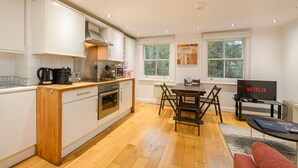 A modern kitchen with white cabinets, wooden countertops, and a dining area with a table and chairs. A television displaying 'NETFLIX' is visible in the background. - The Play Pen - Holborn - by Frankie Says (London)
