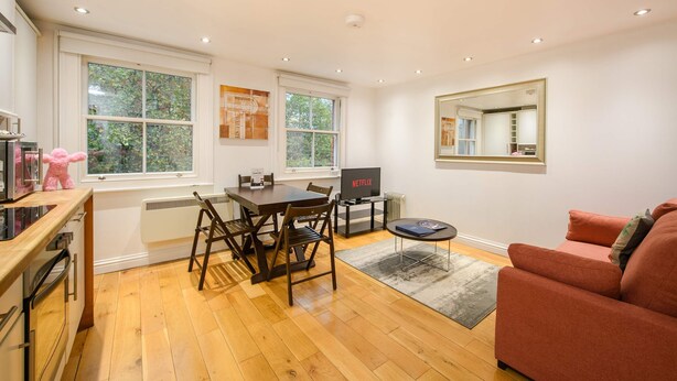 A modern living room with a wooden floor, a red sofa, a dining table with chairs, a TV, and a large window. - The Play Pen - Holborn - by Frankie Says (London)