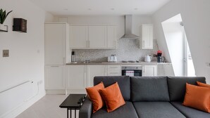 A modern kitchen with white cabinets, a gray sofa with orange cushions, and a small table. - Soul in the City - Fitzrovia - by Frankie Says (London)