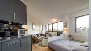 Ein kompaktes Studioapartment mit Küchenzeile, einem Bett, einer Sitzecke mit Stuhl und kleinem Tisch sowie großen Fenstern mit Blick auf die Stadt.
