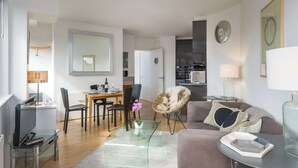 Ein modernes Wohnzimmer mit einem Glastisch, einer Couch, einem Essbereich mit Tisch und Stühlen und einer Küche im Hintergrund.