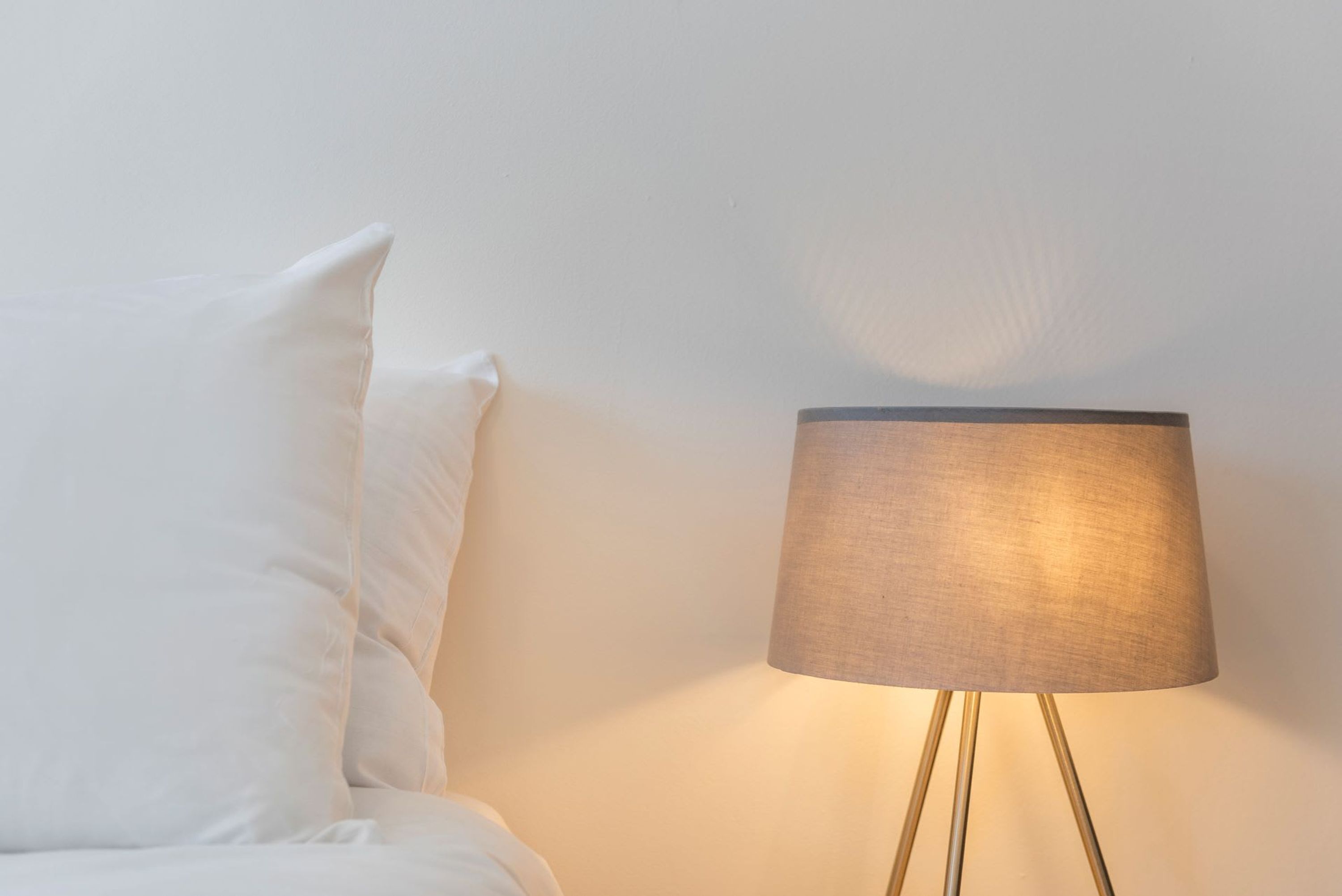 A bed with white bedding and a lamp with a beige shade and gold legs.